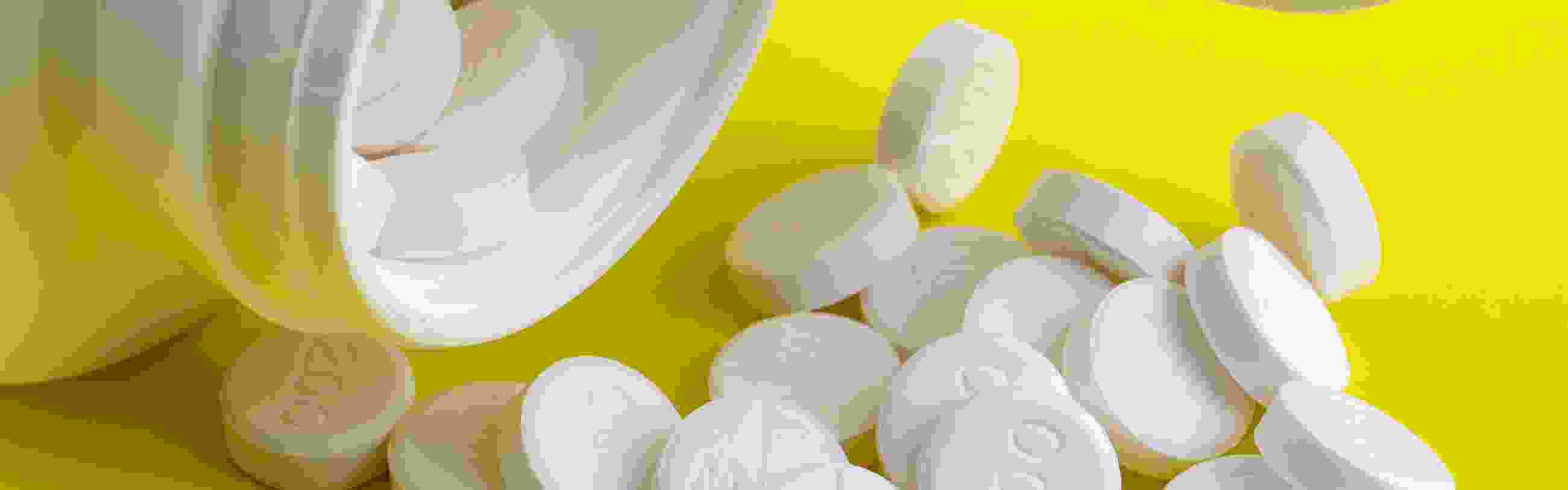 Round white pills spill out of a bottle onto a yellow background