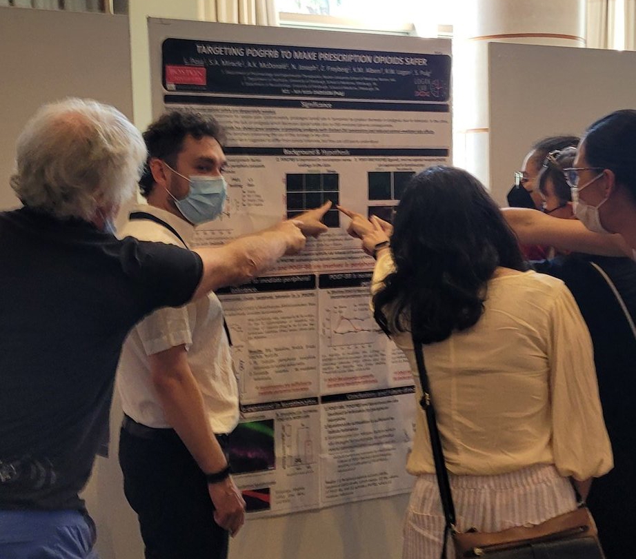 A scientific poster presenter engaging with a small crowd of people, who are pointing at images on the poster. 
