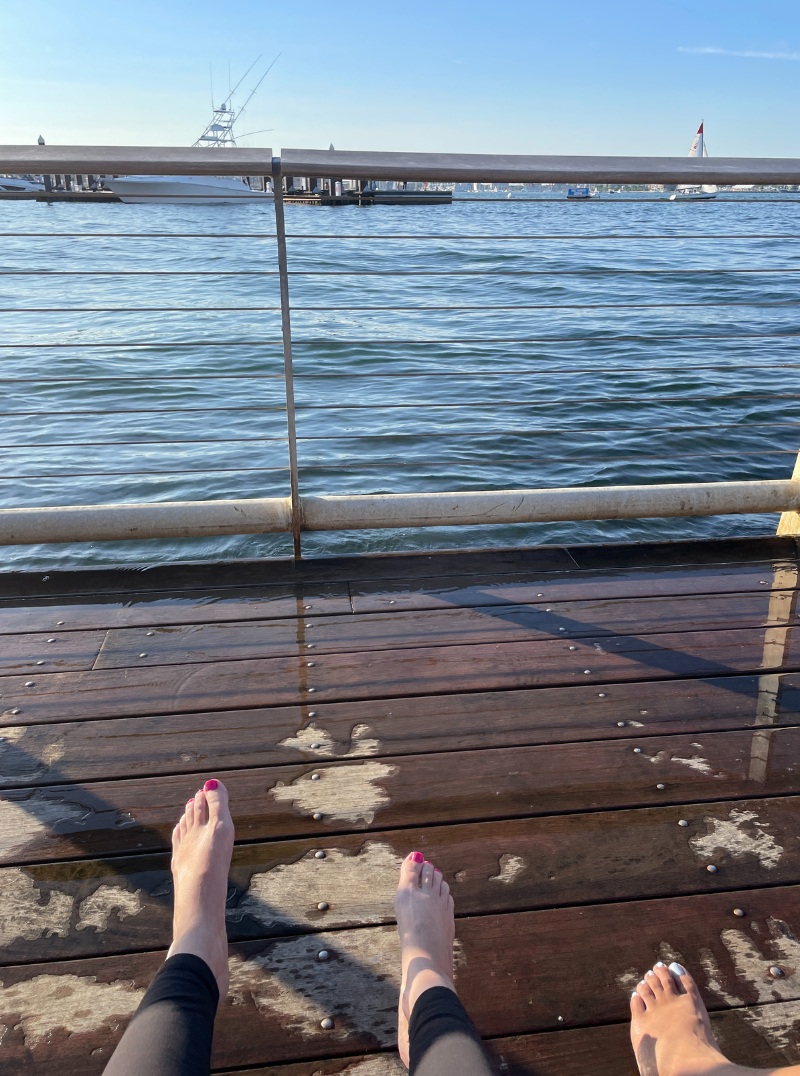 2 people sitting on the dock looking out at the harbor with their lower legs and toes showing