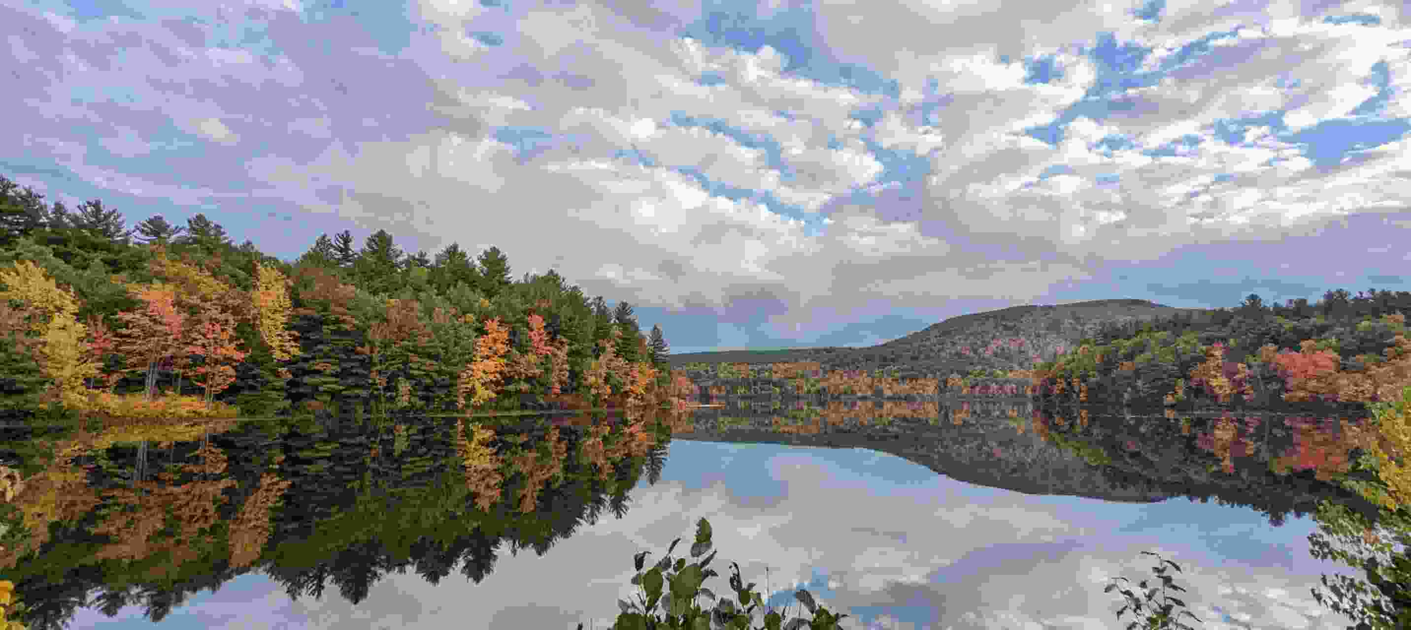 Panoramic view of a lake surrounded by trees with fall foliage and a cloudy blue sky