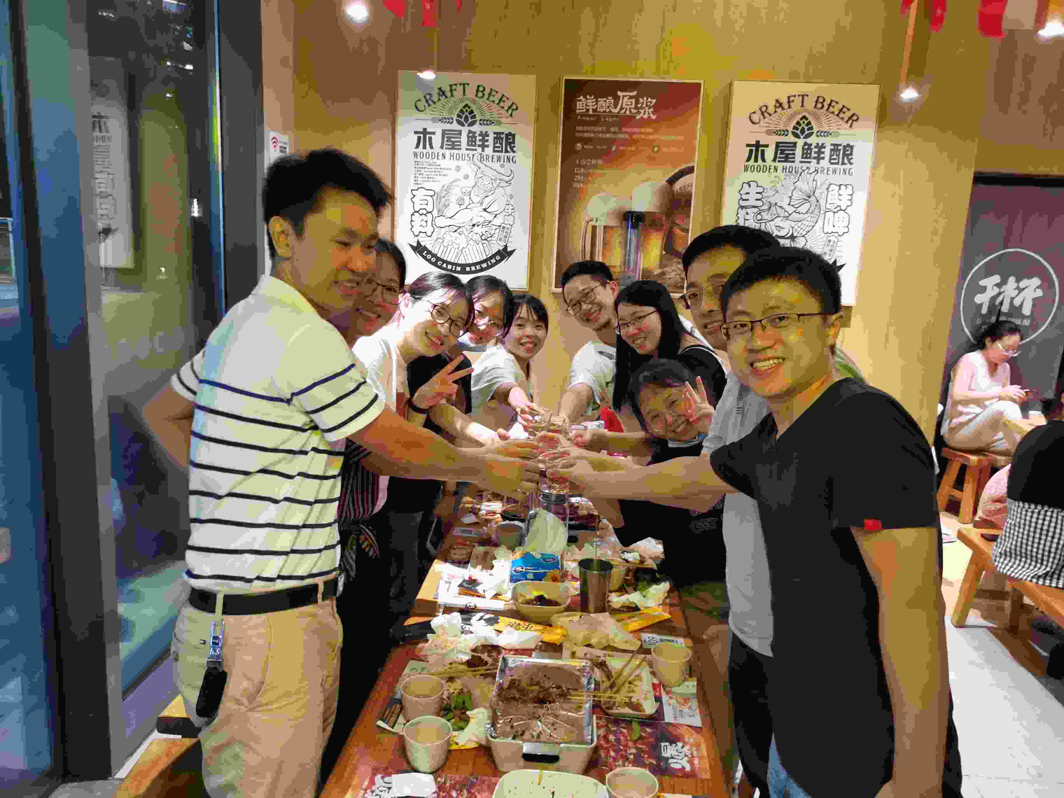 Zhong lab members stand around a table of food, smiling and raising their drinks together over the table's center