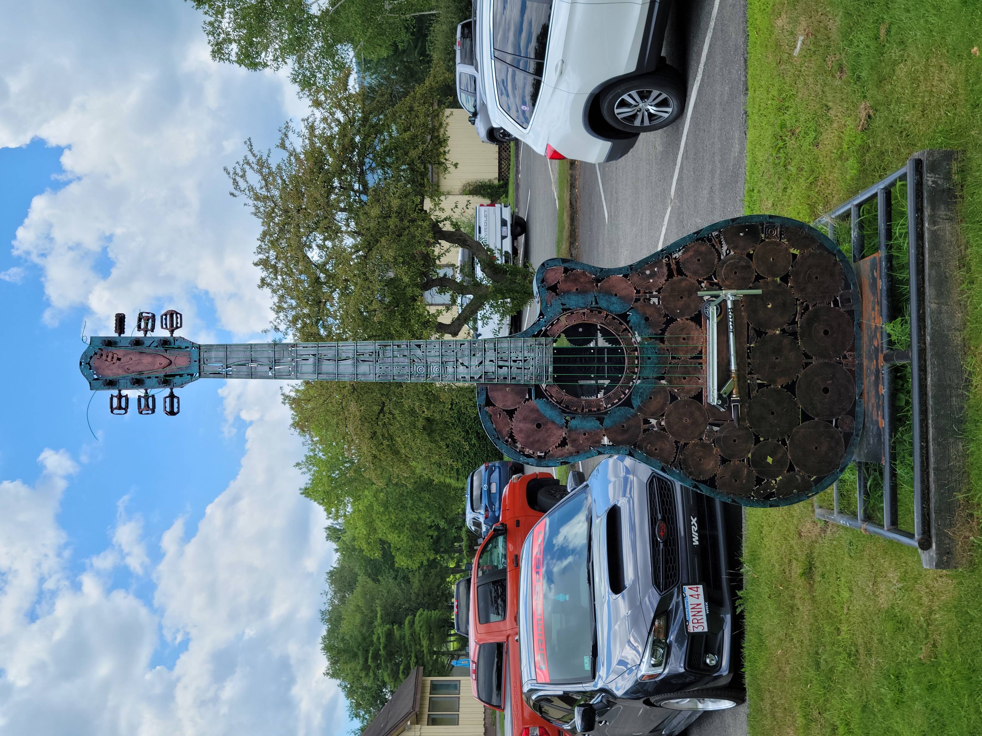 A statue of a guitar made of different metallic materials sits in front of a parking lot full of cars